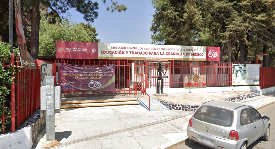 Fotografía en la que se muestra la entrada del plantel, conformada por una reja de color rojo. Sobre la rej<a hay un cartel que dice el nombre del plantel. Frente a la rej<a hay una escalera y una rampa que dan acceso a las instalaciones.